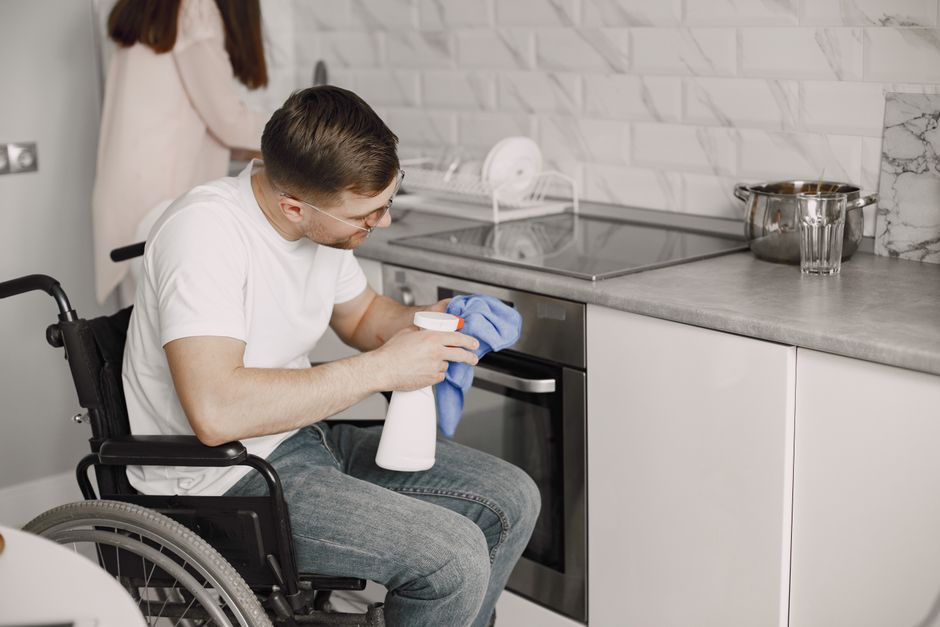 A man using a wheelchair cleans a modern kitchen surface with a spray bottle and cloth.
